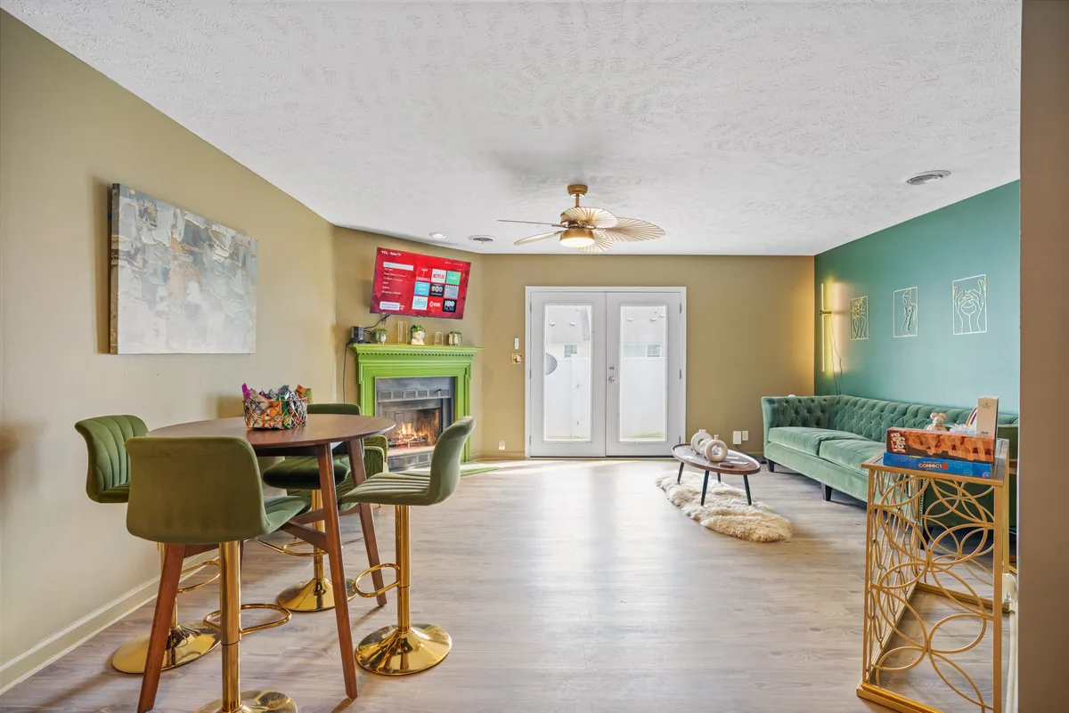 A cozy living area anchored by a green fireplace, a blue velvet sofa, a dining table with high stools, and a TV with streaming options. There are large glass doors providing natural light, and the room is set up to accommodate a group for both meals and relaxation.