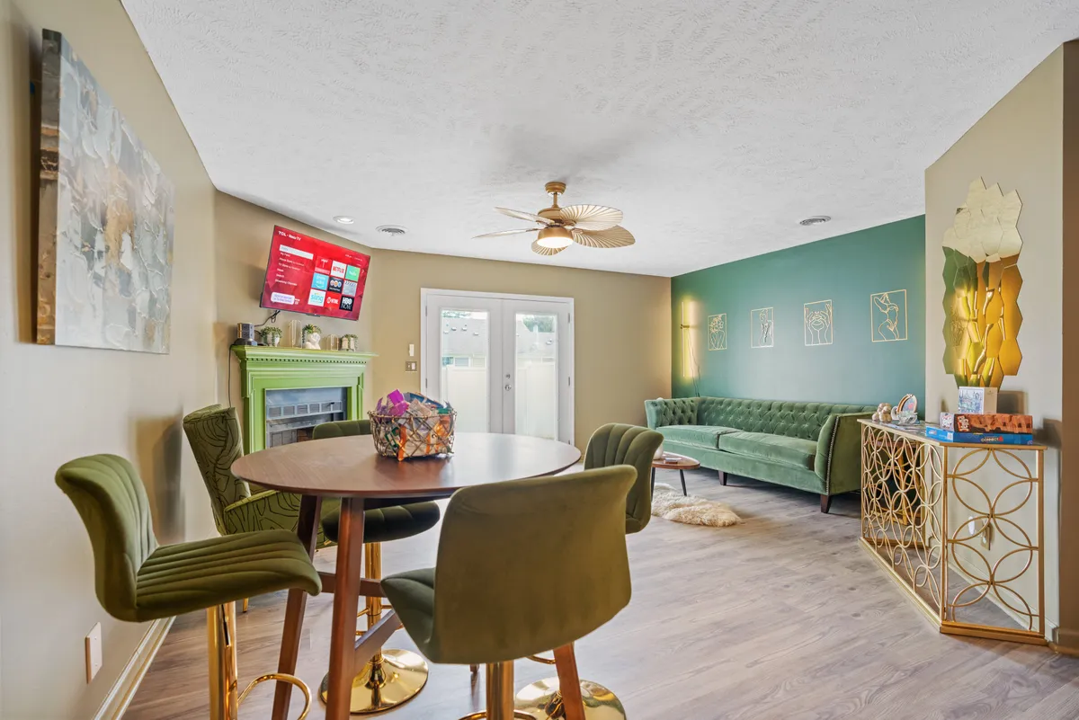 A view of a green velvet sofa paired with a round wooden dining table and a TV displaying streaming content. The golden accent table and colorful decor create a lively yet comfortable ambiance. The room is staged for socializing and entertainment, showing a seamless flow between spaces.