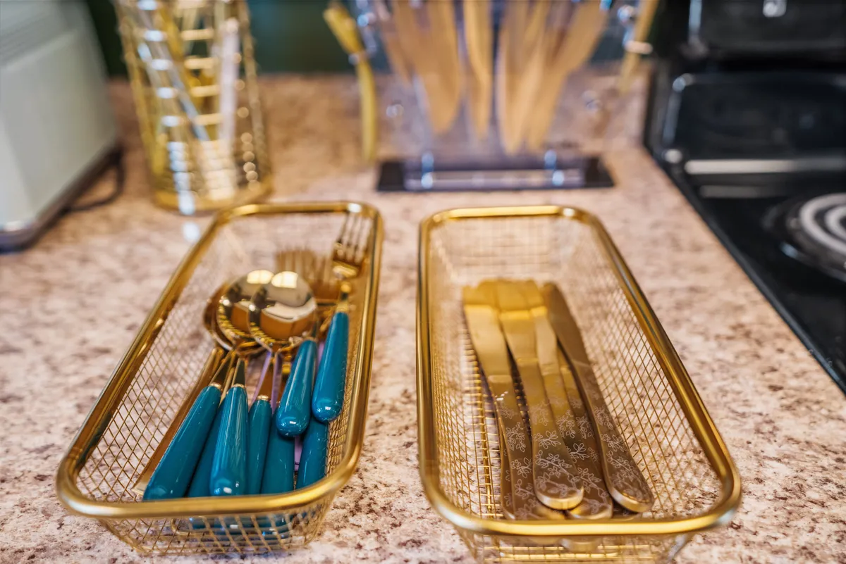 A close-up of a stylish kitchen countertop featuring golden utensils, teal-handled cutlery, and textured surfaces. The scene is set for meal preparation and entertaining, with design touches that add a sense of modern elegance and warmth.