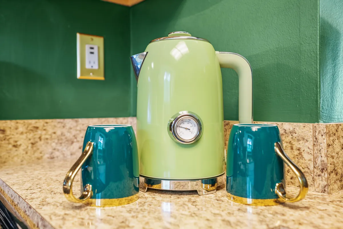 A cheerful countertop vignette featuring a green kettle, teal mugs, and a granite surface. The setup suggests a cozy moment of brewing tea or coffee, perfect for morning breakfasts or late-night chats around the dining table.