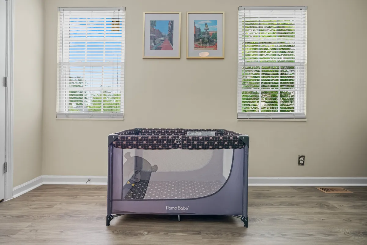 A playpen is situated in a sunlit room with windows and framed pictures on the wall, signaling a welcoming, child-friendly environment. The space is designed for both play and family interaction.