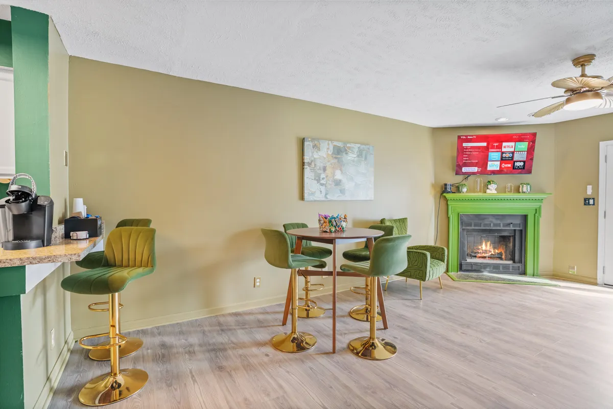 This image features a stylish open-concept living area with a green fireplace, round dining table, bar stools, and a TV with streaming services. The furniture is arranged to promote social interaction and comfort, and the warm color palette adds to the inviting atmosphere. The fireplace is a focal point, and the modern touches support group gatherings.