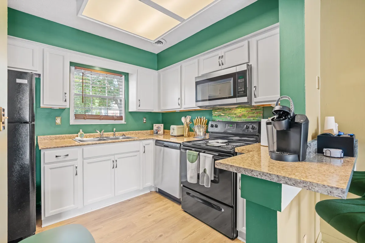 This image captures a vibrant kitchen scene with a green accent wall, modern appliances, a clean countertop, and a bar seating area. Natural light pours in through the window, brightening the space and emphasizing its cheerful, sociable vibe. The bar seating and open layout suggest a seamless flow between cooking and entertaining.